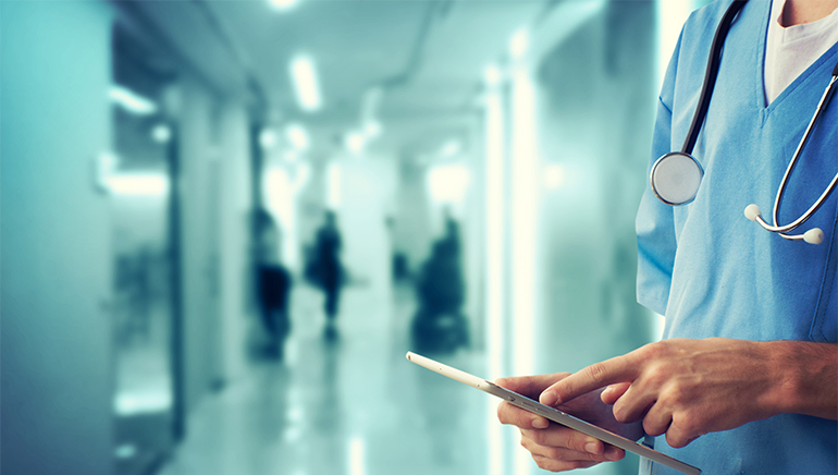photo - closeup of a person wearing medical scrubs and a stethoscope in a hospital hallway using a tablet device
